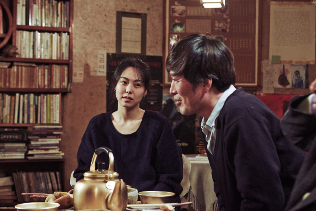 영화 지금은 맞고 그때는 틀리다 스틸컷, 책장이 늘어선 술집에서 노란 막걸리 주전자를 앞에 두고 마주 앉은 두 사람