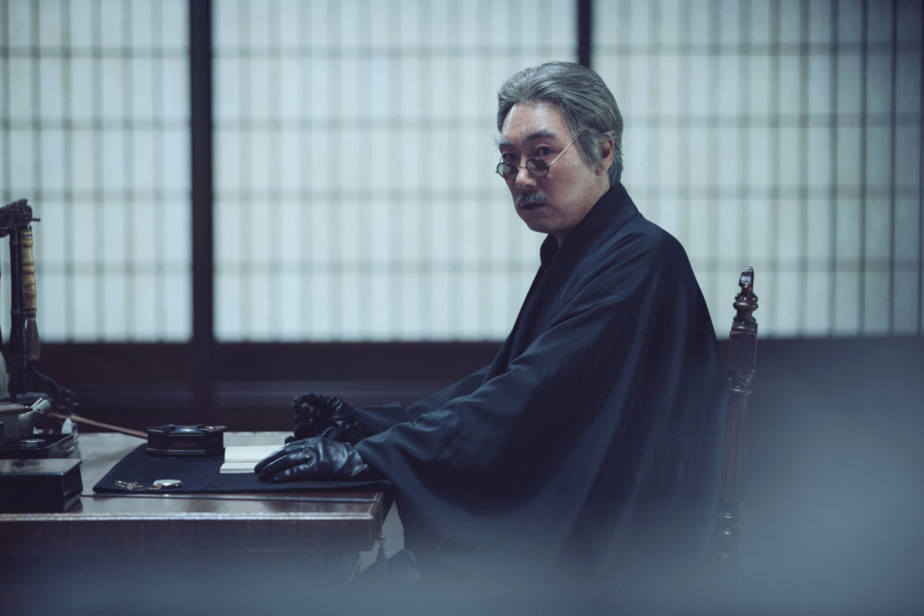 영화 아가씨 스틸컷, 장지문 앞 책상에 검은 장갑을 끼고 앉아 돌아보는 코우즈키(조진웅)